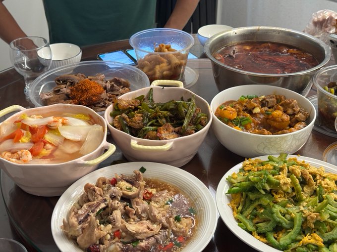 A spread of home-cooked Chinese dishes—including spicy hot pot, stir-fried bitter melon with eggs, braised meat with potatoes, and other hearty plates—is laid out on a table, shared among coworkers after a chest workout. The post thanks a colleague nicknamed "五老师" for the excellent cooking while joking about daily team-building meals at CDC.