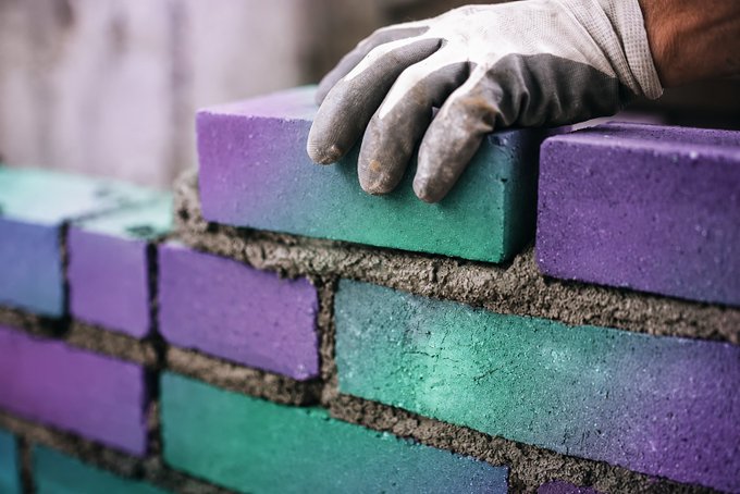 Close-up photo of a gloved hand placing a purple and teal gradient brick onto a wall of colorful bricks being laid with mortar.