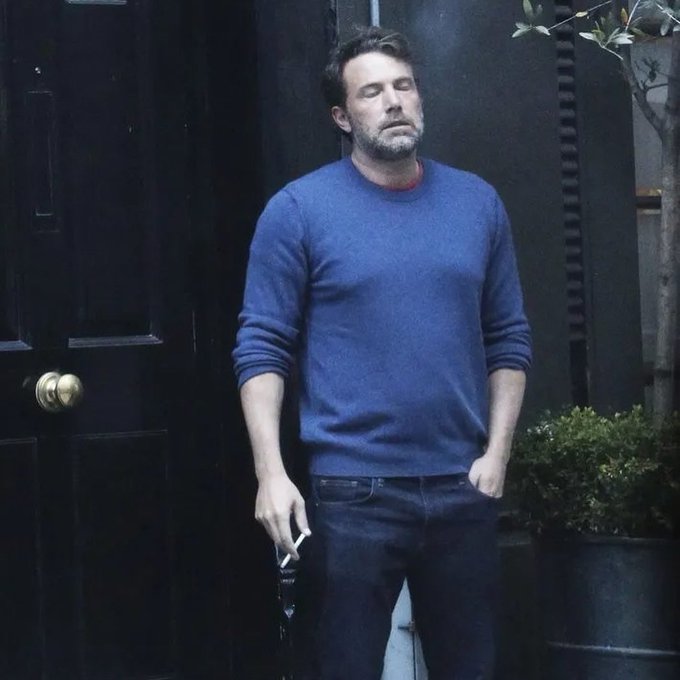 A man in a blue sweater and jeans standing by a black door with eyes closed, holding sunglasses.