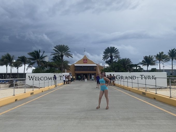 A person poses on a pier in front of a 'WELCOME TO GRAND TURK' sign, with a beach, palm trees, and an orange building visible in the background under a cloudy sky.