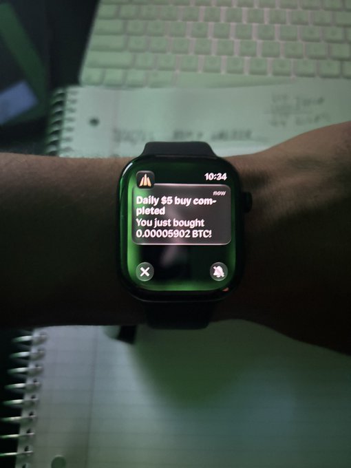 A smartwatch on a wrist displays a notification: 'Daily $5 buy completed. You just bought 0.00005902 BTC,' positioned over a keyboard and notebook.