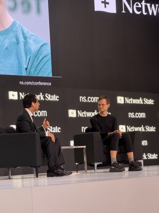 Two men sit in armchairs on stage at a 'Network State' conference, with 'ns.com/conference' visible on the backdrop behind them.