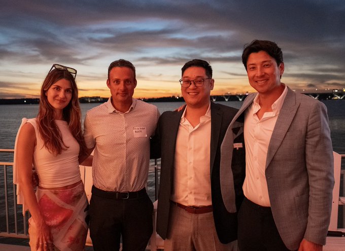 Four people smile for a photo at sunset by the water, with a bridge visible in the distant background.