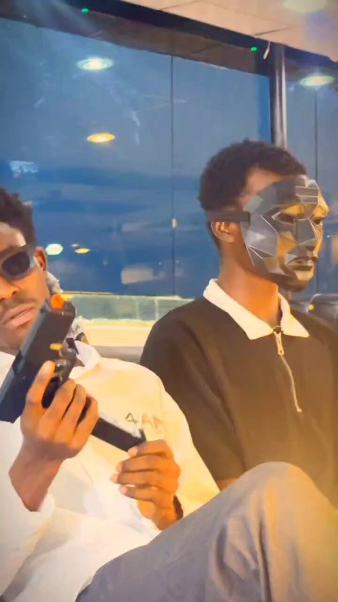 The image features two men sitting next to each other, both wearing masks that cover their faces. One of them is holding a gun while the other one has a video camera attached to his face. The man with the mask appears to be playing a game on the video camera, and they are both seated in chairs.