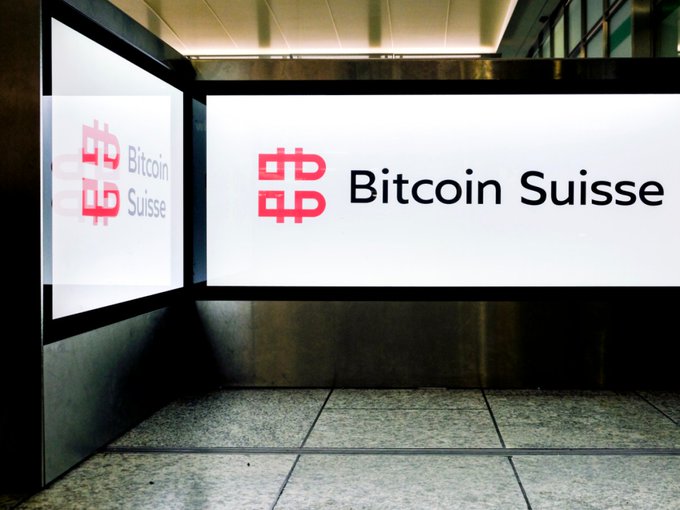 A brightly lit corner displays the "Bitcoin Suisse" company name in red and black text, with a second sign on the left side, in a building that the post says is the cryptocurrency firm's headquarters in Zug, Switzerland, where it is considering cutting up to 60 of its 120 local jobs amid a restructuring that will move back-office and software operations to an existing site.