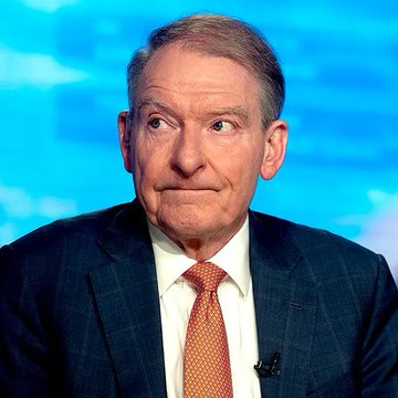 A man in a dark blue suit, white shirt, and orange patterned tie sits in front of a blue cloudy backdrop, likely during a news interview or broadcast.