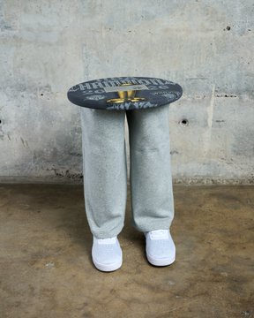 A surreal stool sculpture with fabric-covered human legs and white sneakers supporting a blue printed seat that reads "TRUMP" sits against a concrete wall, embodying the absurd art piece being mocked in the post about SuperRare promoting a table with human legs from a little-followed creator.