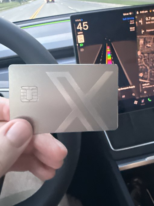 A hand holding a metallic card with an 'X' logo inside a Tesla, with the dashboard showing a speed of 45 MPH and navigation.