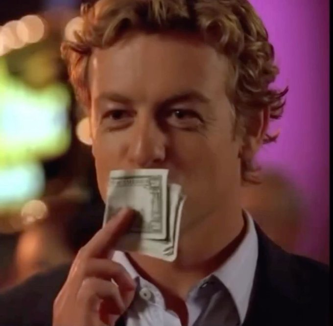 A curly-haired man in a dark jacket holding cash bills near his mouth against a purple-lit background, appearing to be from a movie scene.