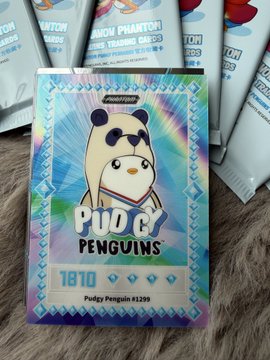 A holographic Pudgy Penguins trading card (#1810, Pudgy Penguin #1299) resting on a fuzzy surface, with Phantom Phantom trading cards visible in the background.