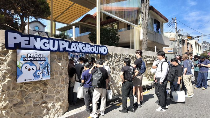 A photograph of a crowd of people gathered outside a venue called 'PENGU PLAYGROUND' on a sunny day, with a sign featuring a penguin mascot.