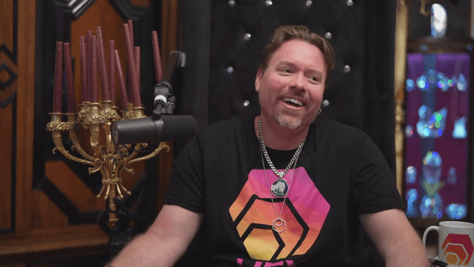A smiling man with a goatee sits in a studio setting, wearing a black t-shirt with a colorful graphic, a necklace, with a decorative candelabra behind him.