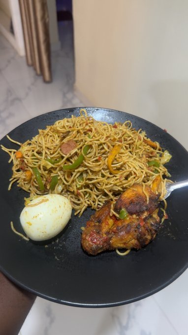 A black plate containing a meal of stir-fried noodles with vegetables and sausage, a hard-boiled egg, and a piece of grilled chicken.