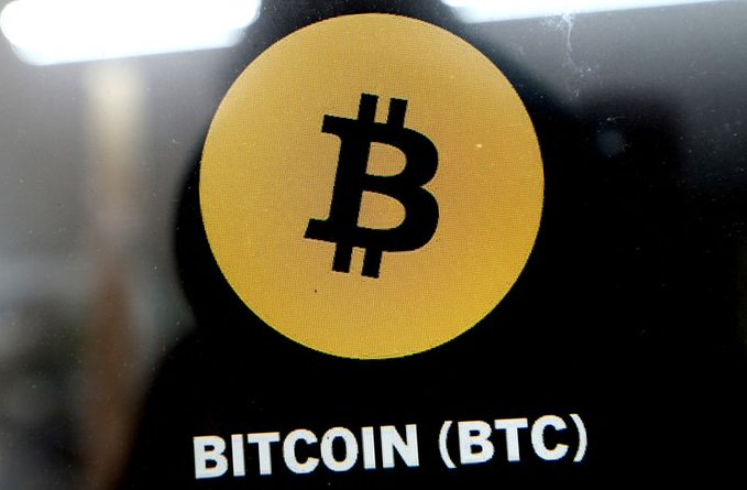A close-up of a yellow Bitcoin logo with the text 'BITCOIN (BTC)' displayed on a screen.
