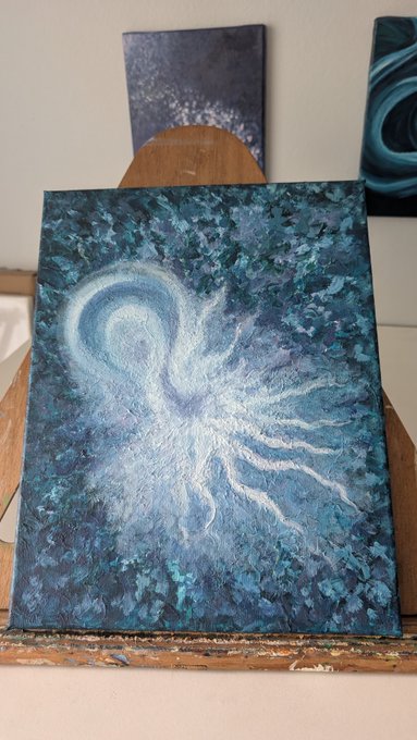 A textured acrylic painting featuring a luminous white angel figure with flowing wings against a deep blue and teal mottled background, displayed on a wooden easel.