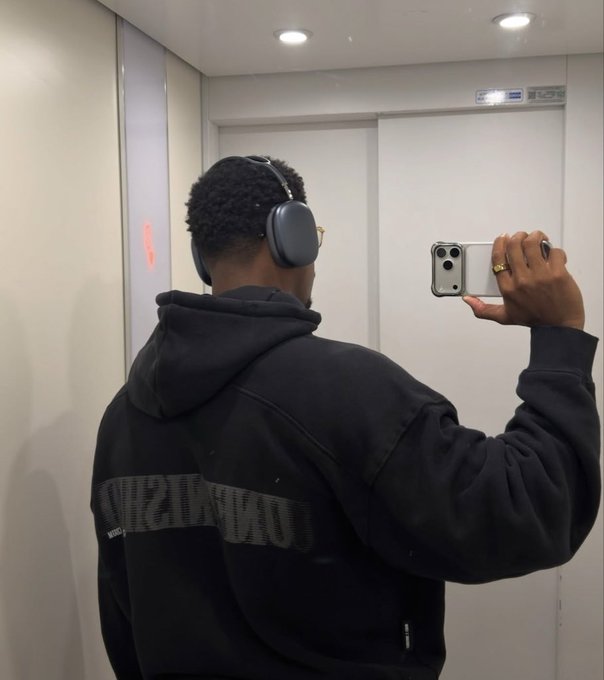 A mirror selfie of a person wearing a black hoodie, headphones, and glasses, holding an iPhone to take the photo, with a gold ring visible on their finger.