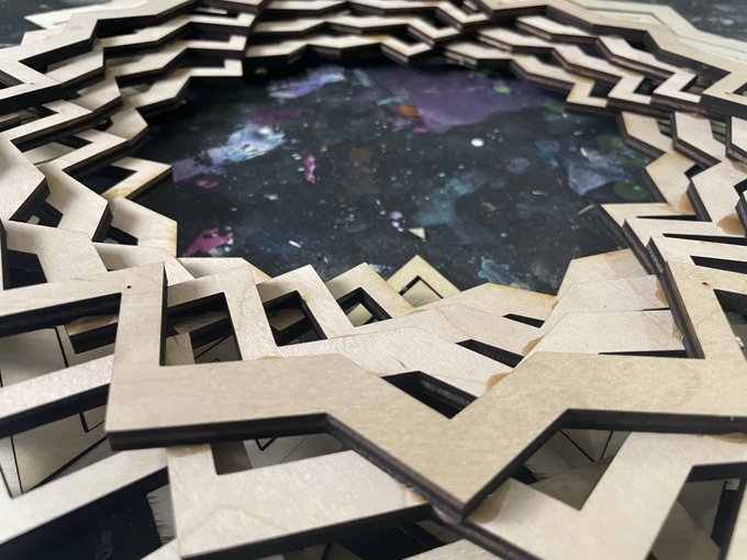 A close-up photograph of a decorative wooden object featuring an intricate, interlocking geometric star pattern cut out of MDF or similar wood, sitting on a dark, paint-splattered surface.