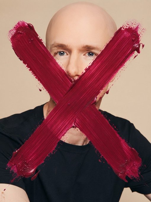 A bald man in a black shirt with a large red paint X painted across his face.