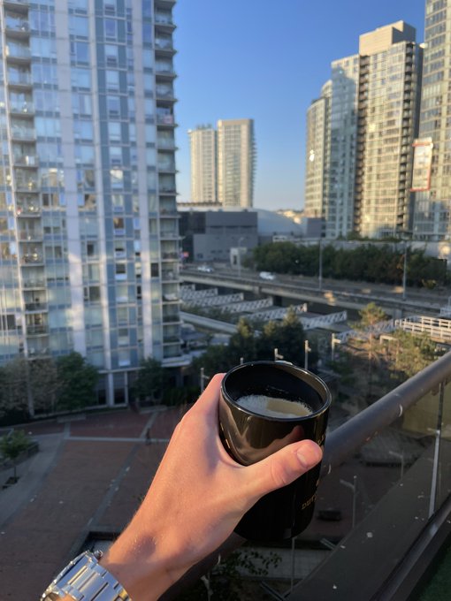 A hand holds a black mug against a sunny urban skyline of glass high-rises, captioned "Gmgm" — a casual, content mood as if enjoying a quiet morning coffee overlooking the city.
