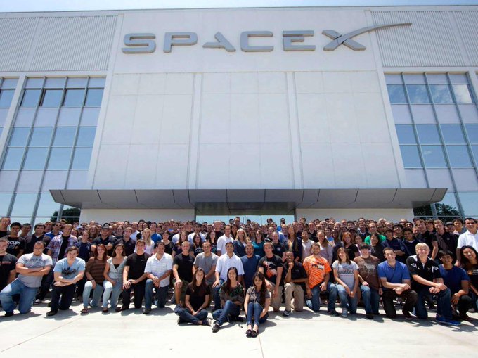 The image features a large group of people posing for a photo outside of a white building, which appears to be a company or office space. The individuals are standing close together, creating an impression of unity and camaraderie among them. The photo captures this moment in time, showcasing the diverse group of people gathered at the location.
