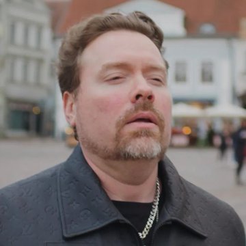 A close-up photograph shows Richard Heart, the founder of HEX, PulseChain, and PulseX cryptocurrencies, standing outdoors in what appears to be a European city square with historic architecture in the background. He has curly reddish-brown hair, a goatee, and is wearing a dark jacket with a silver chain necklace, his eyes nearly closed as the image appears to be a still frame from a video.