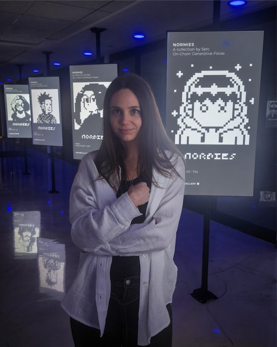 A young woman stands in a dark gallery space between illuminated displays featuring pixelated "NORMIES" characters, with a large screen behind her showing one of the on-chain generative faces.