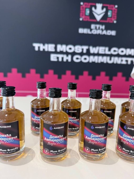 Mini bottles of "Домаћа Амбајеровка Југа 2020" Plum Brandy by Ambire are lined up on a table at an ETH Belgrade event, featuring the slogan "THE MOST WELCOME ETH COMMUNITY" in the background. The post's mention of "Ambirakijca" is a playful nod to the Ambire brand of plum brandy pictured, hinting that a few of these shots might help attendees blend into the crowd.