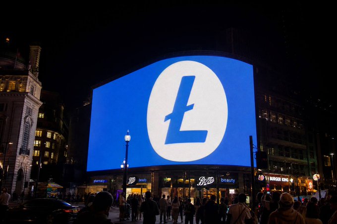 A nighttime photograph of a large illuminated digital billboard displaying a stylized white 'L' logo on a blue background at Piccadilly Circus, with crowds gathered below.