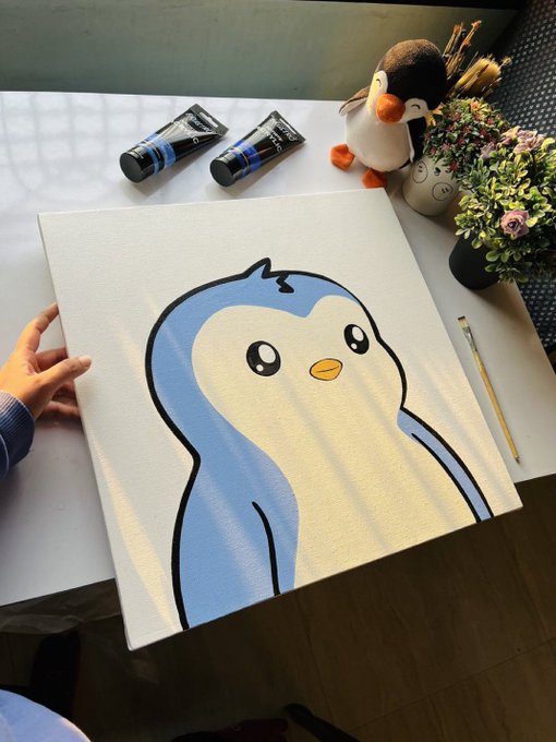 A hand holds a small canvas painting of a cute blue and white cartoon penguin, displayed on a table next to acrylic paint tubes, a brush, and a stuffed penguin toy.