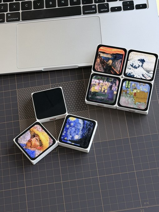 Several small, square, pixelated display devices showcase various digital artworks—including images resembling Vincent van Gogh's "Starry Night" and classical paintings—arranged next to a MacBook laptop on a cutting mat. The image visually represents the concept of "Hardware NFTs" by displaying physical gadgets that display digital/pixel art collectibles.