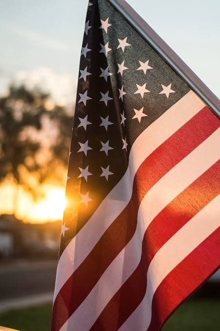 An American flag is shown backlit by a golden sunset, with no visible text. The image symbolizes the patriotic backdrop of the CLARITY Act's partisan battle over U.S. digital asset regulation.