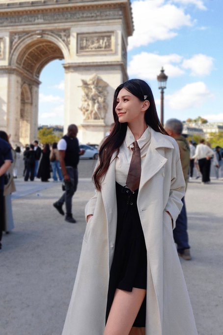 A young woman with long dark hair stands in front of the Arc de Triomphe in Paris, wearing a black dress under a long cream-colored coat.