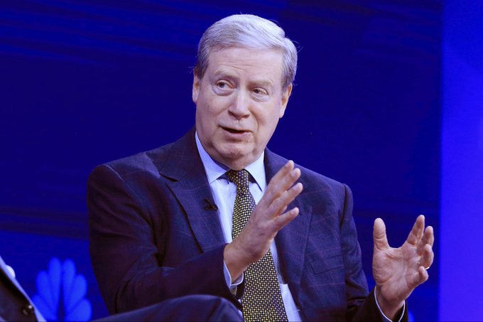 A man in a dark suit and patterned tie is speaking on stage, gesturing with his hands against a blue background with partial logos visible at the bottom. The post references cryptocurrency tickers $TAO, $TIG, and $TIBBIR, which appear unrelated to the image of the speaker shown.