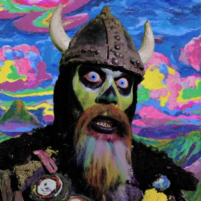A surreal, colorful painted portrait of a viking-like figure with a horned helmet, green skin, blue eyes, and a multicolored beard against a psychedelic landscape.