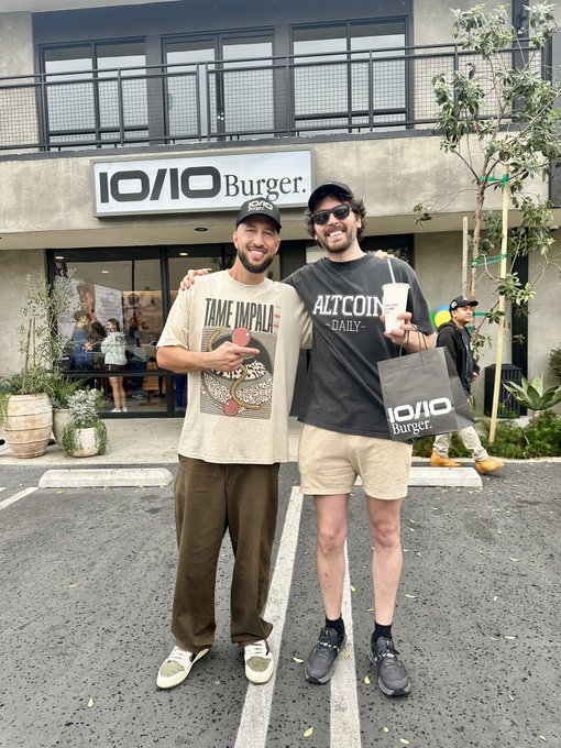 Two friends pose outside the 10/10 Burger restaurant, with one wearing an "Altcoin Daily" t-shirt and holding a branded 10/10 burger bag, celebrating their visit on the numerically perfect date of 10/10.