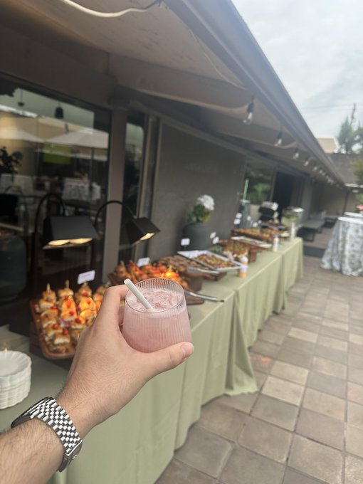 A first-person view of someone holding a pink cocktail at an outdoor catered event with a long green-skirted buffet table displaying various appetizers.
