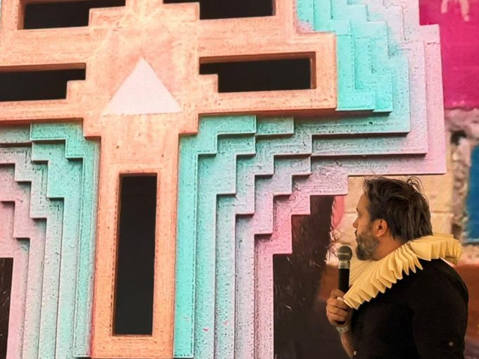 The image features a man standing next to a large, colorful, and intricate sculpture made of many small panels or blocks stacked together. He appears to be giving a speech or presentation while holding a microphone. The sculpture itself has a unique design with various colors and patterns that make it visually striking. The post in the image reads "Epic Gm", which could potentially be related to the artwork, such as being part of an art exhibition or showcasing the artist's work.

The man is wearing a yellow scarf around his neck, adding a pop of color to the scene. The combination of the colorful sculpture and the man speaking into the microphone creates an interesting contrast between the artistic and the spoken word.