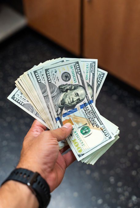 A hand wearing a black smartwatch holds a fan of multiple US $100 bills.