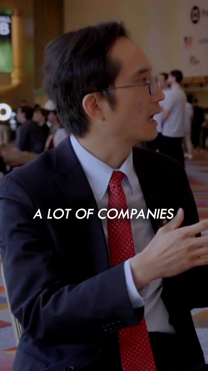 The image features a man wearing glasses, a suit, and a red tie who appears to be talking to someone else. The background shows several people sitting at tables or standing around, suggesting that they are gathered for an event or meeting. There is also text in the image that reads "A Lot of Companies", which could potentially be related to the man's profession as a businessman or the nature of the gathering itself.