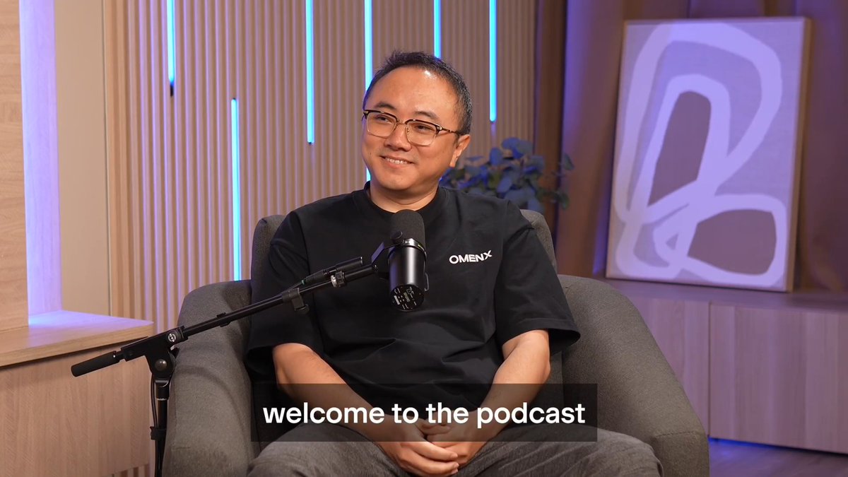 A man wearing glasses and a black OMENX t-shirt sits in a chair with a microphone in front of him, with the caption "welcome to the podcast" displayed.