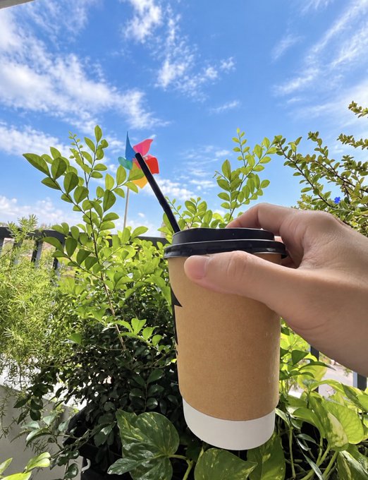 A hand holding a brown paper coffee cup outdoors, with a colorful pinwheel and green foliage visible against a blue sky.
