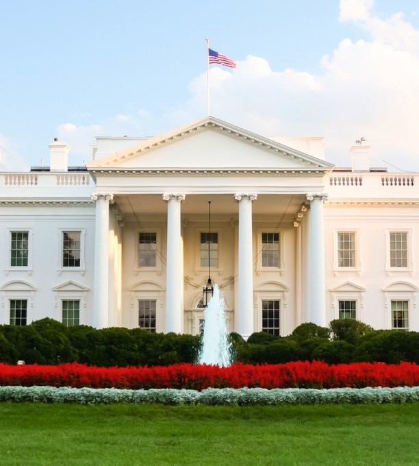 A daytime photograph of the North Portico of the White House, featuring its white columns, an American flag flying on top, and a manicured garden with red flowers in the foreground.