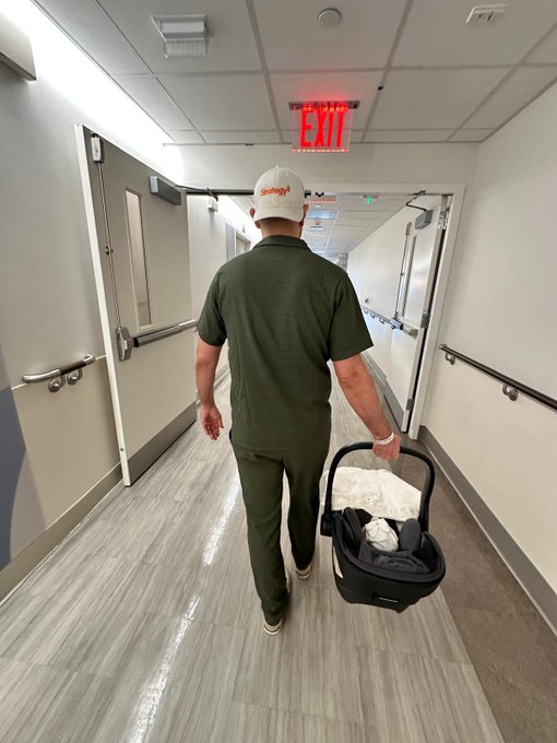 A man wearing a white cap, olive green polo shirt and matching pants walks down a hospital corridor carrying a black infant car seat, passing under a red illuminated EXIT sign.