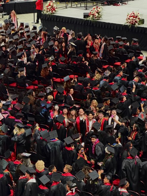 A large crowd of graduates in black caps and gowns, many adorned with colorful leis, gather and celebrate at a commencement ceremony. The image ties to "Game of Loans" by depicting the culmination of years of educational investment, symbolizing the financial reality many graduates face as they transition from academic life into repaying their student debt.