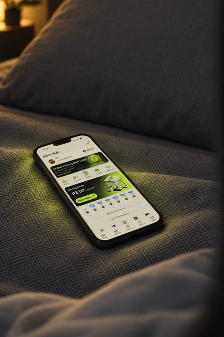 A smartphone displaying the Binance app interface rests on a bed with gray textured bedding and pillows.