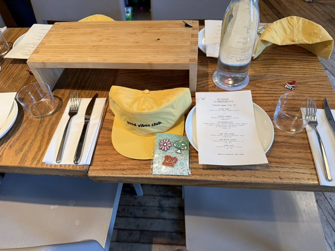 A wooden restaurant table setting with 'good vibes club' branded yellow caps, wine glasses, silverware, a menu, and a water bottle.