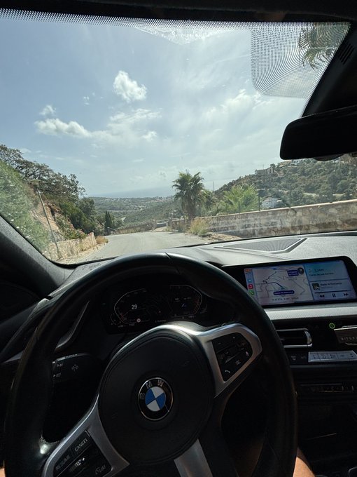 A first-person view from inside a BMW driving down a sunny road, with the steering wheel and dashboard navigation visible.