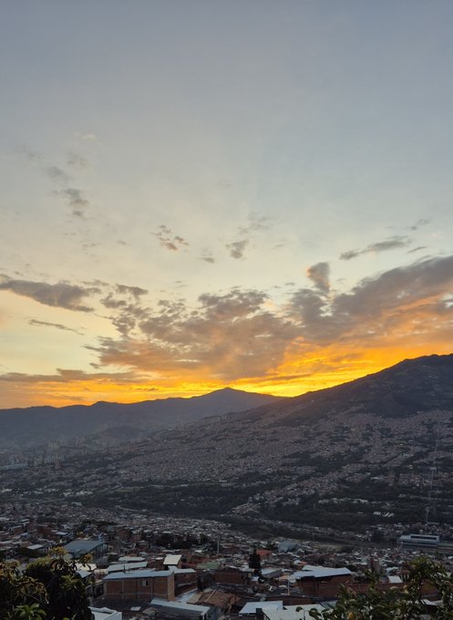 A scenic sunset over a sprawling city nestled in a valley, with mountains in the distance and warm orange and yellow hues filling the cloudy sky above densely packed buildings.