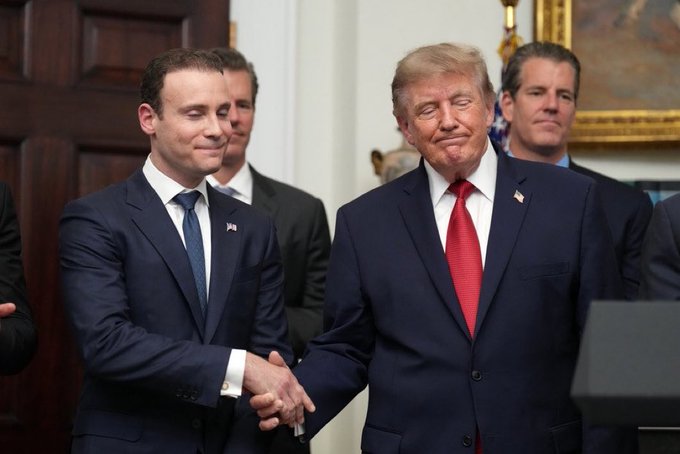 A photograph showing a handshake between a man in a dark suit with a blue tie (with an American flag pin) and Donald Trump, who is wearing a dark suit with a red tie, in what appears to be the White House.