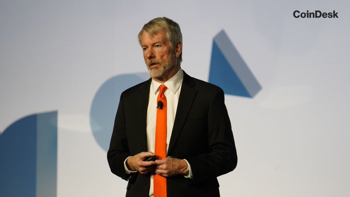 Michael Saylor, MicroStrategy's executive chairman, speaks at a CoinDesk event while wearing a black suit and bright orange tie, with the "CoinDesk" logo visible in the upper right corner of the image. His appearance accompanies the news of his statement that "Bitcoin doesn't need CLARITY. America needs clarity," made amid the CLARITY Act's failure to receive a Senate vote before the August recess.